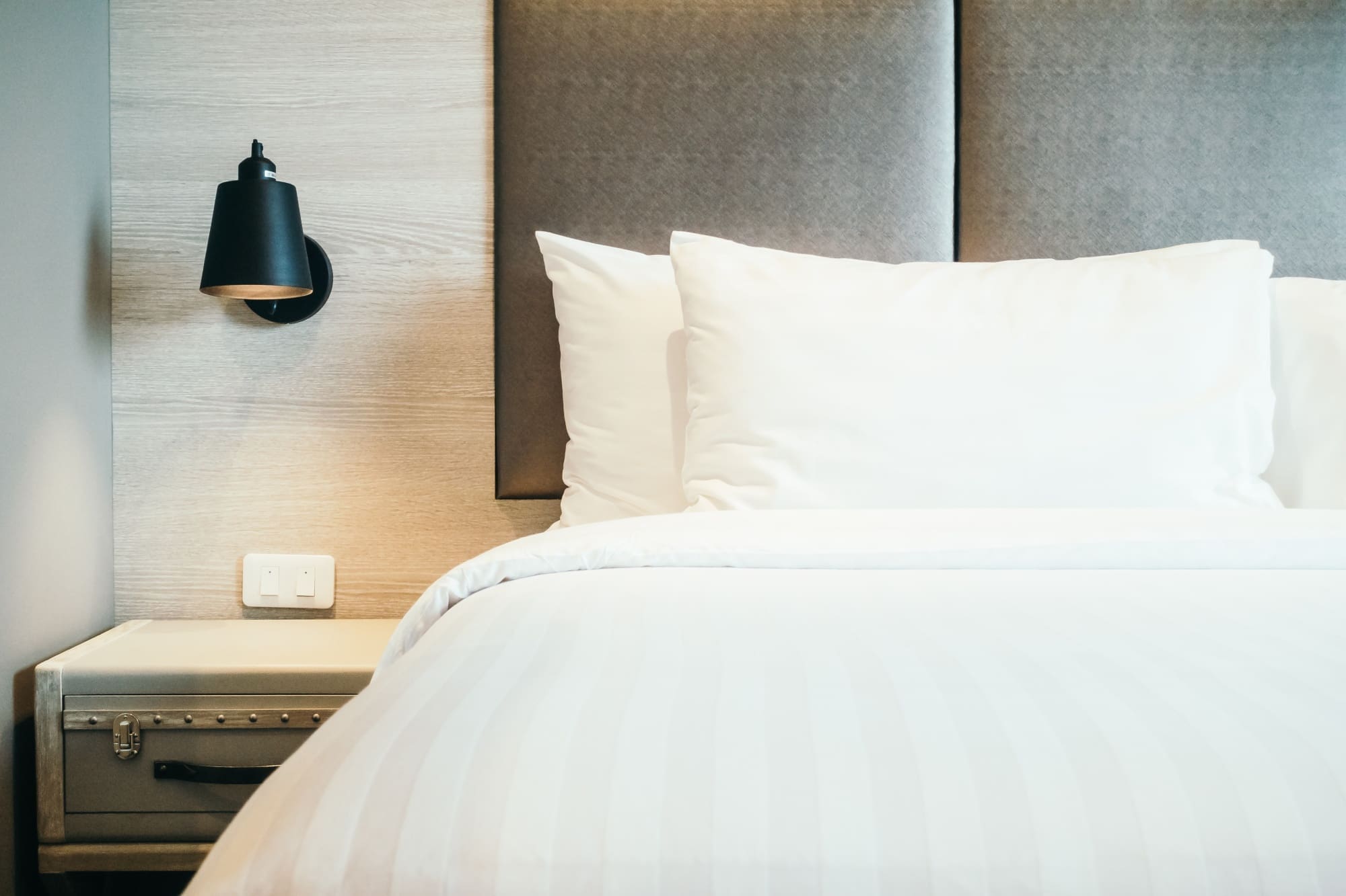 A neatly made bed with white linens, two pillows, a padded headboard, a bedside table, and a black wall-mounted lamp.